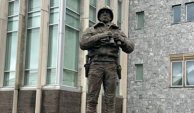 The photo shows a bronze statue of a soldier holding binoculars, standing in front of a modern building with large windows and a stone facade.