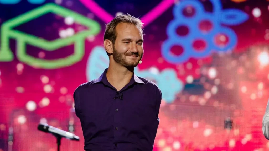 A man in a purple shirt is standing on a stage with a colorful background, smiling and looking to his right.