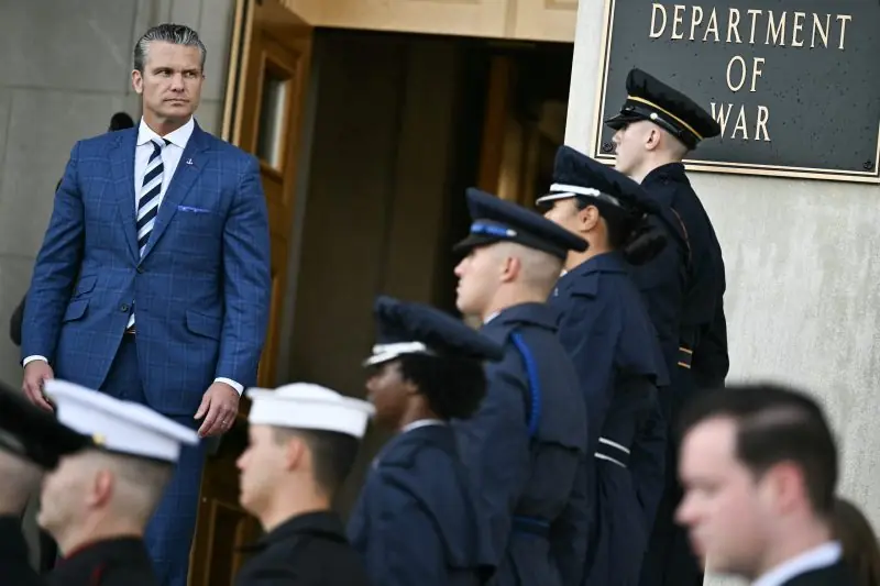 A man in a blue suit stands in front of a group of uniformed personnel in front of the Department of War.