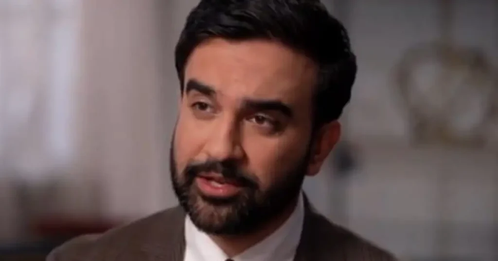 The image shows a man in a suit and tie, speaking in what appears to be a formal or professional setting.