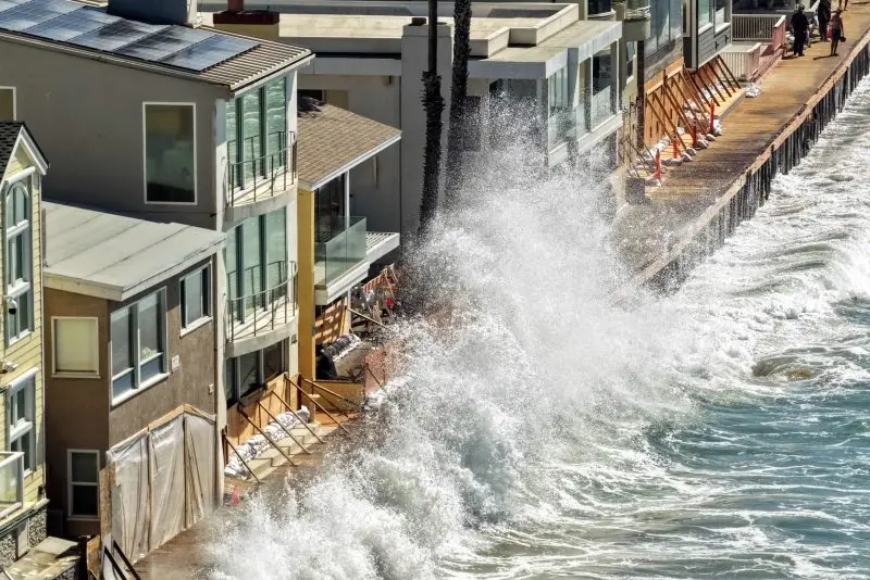 A powerful wave crashes against a coastal structure, threatening nearby buildings and infrastructure.