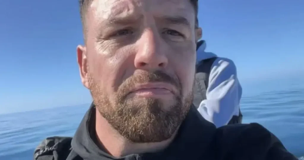 The image shows a person with a beard and a jacket, standing on a boat with the ocean and sky in the background.