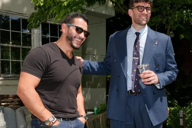 Two men are seen smiling and interacting in an outdoor setting, with one dressed in a suit and the other in casual attire.