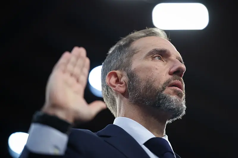 A man in a suit and tie is seen from the side, raising his hand in a gesture of acknowledgment or applause.