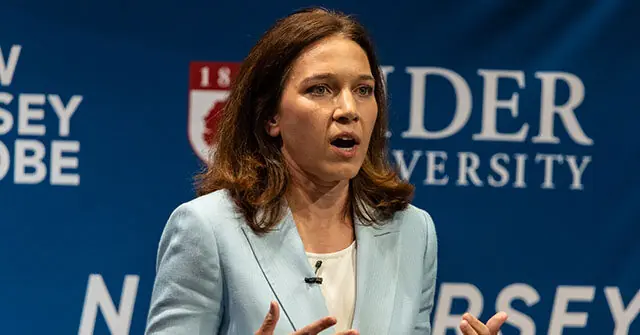 A woman is speaking at a podium in front of a backdrop featuring the "New Jersey" and "Rider University" logos.