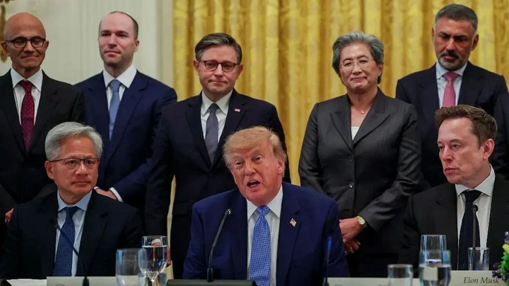 The image shows a formal meeting with a group of individuals in suits, including a man seated at a table with a microphone, addressing the group.