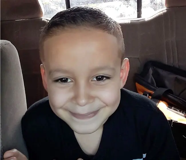 A young boy smiles brightly while sitting in the back seat of a car.