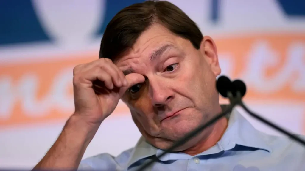 The image shows a man in a light blue shirt, appearing to be in a serious or contemplative mood, with his hand near his face.