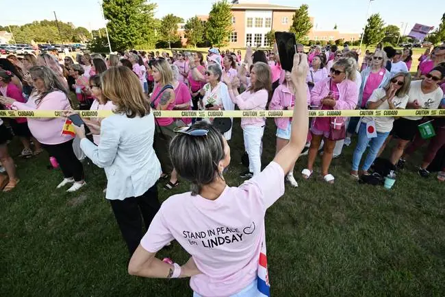 A group of people wearing pink shirts gather outdoors, participating in a peaceful event, with one person raising their hand in a gesture of solidarity.
