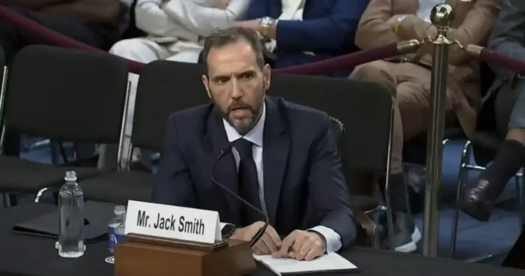 The image shows a man in a suit seated at a table with a nameplate that reads "Mr. Jack Smith," indicating he is likely participating in a formal meeting or hearing.