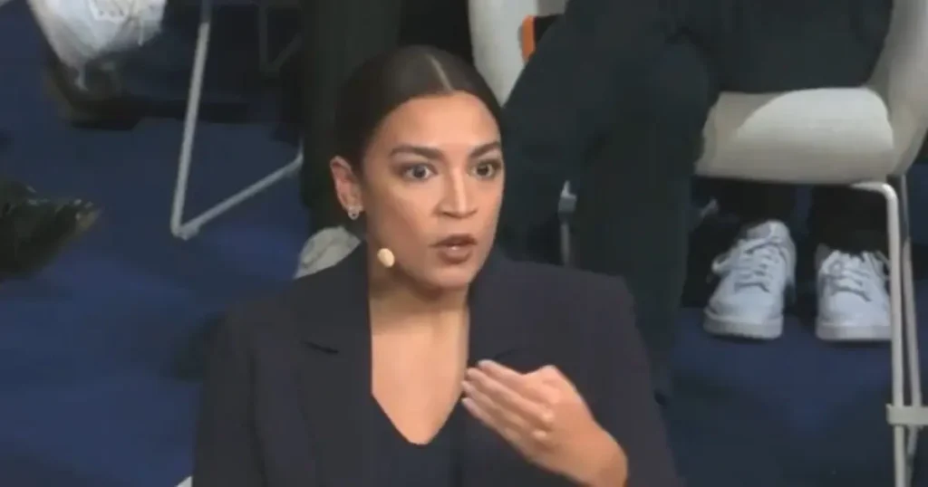 A woman in a black blazer is speaking, with her hand raised, in what appears to be a formal setting.