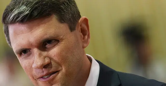 The image shows a close-up of a man in a suit, appearing to be in a formal or professional setting.