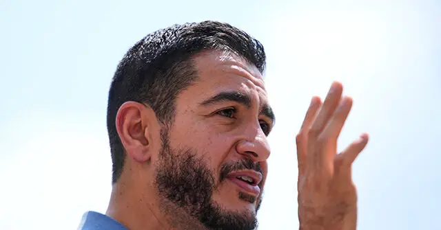 The image shows a man with a beard and short hair, gesturing with his right hand while speaking outdoors under a clear sky.