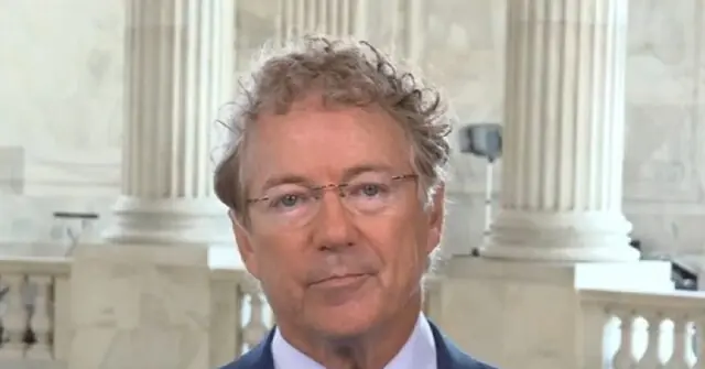 The image shows a man in a suit and tie, wearing glasses, standing in front of a backdrop of classical columns, likely in a formal or governmental setting.