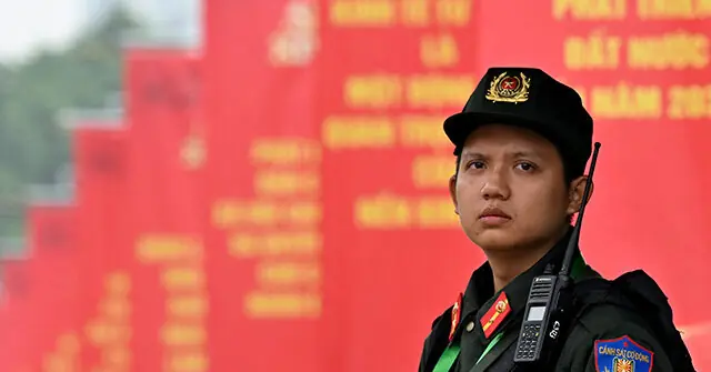 A uniformed officer stands in front of a red backdrop with yellow text, likely at a public event or ceremony.