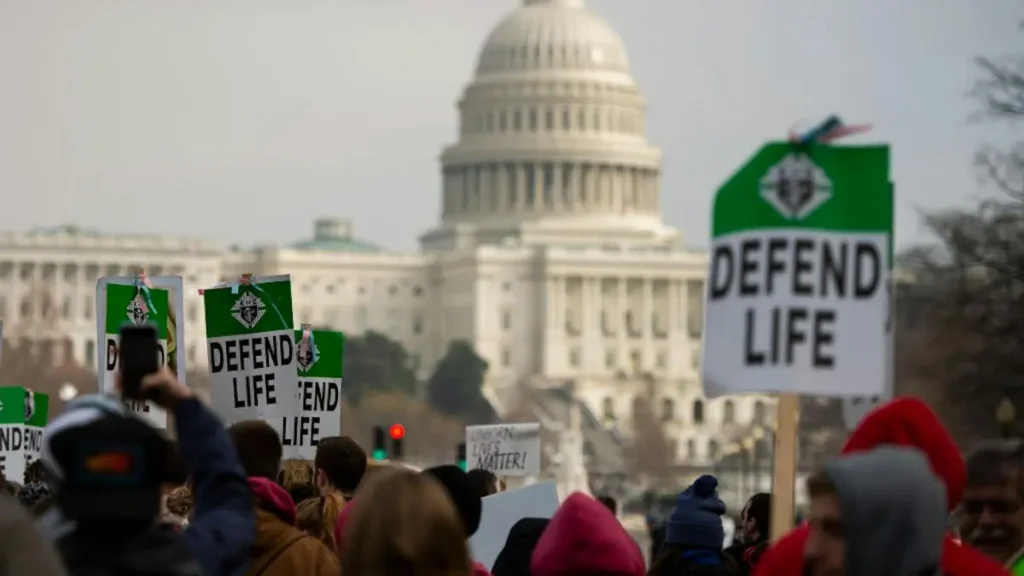 A crowd gathers in front of the U.S. Capitol building, holding signs that read "Defend Life," advocating for pro-life causes.