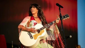 A musician performs on stage, holding a white guitar with intricate designs, while wearing a colorful outfit and a red shawl.