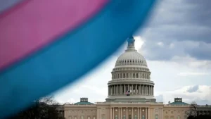 The image shows the U.S. Capitol building with a transgender pride flag in the foreground, symbolizing LGBTQ+ rights and visibility.
