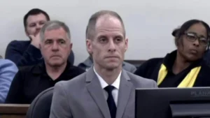 The image shows a group of people seated in what appears to be a courtroom setting, with one individual in the foreground wearing a suit and tie.