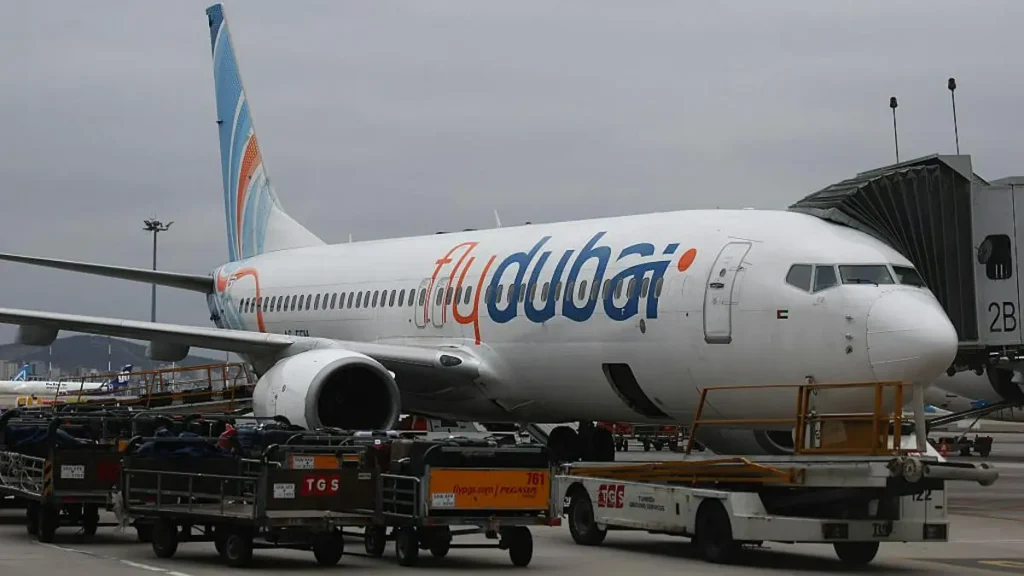A FlyDubai aircraft is parked at an airport gate, with luggage carts and a jet bridge connected to the plane.