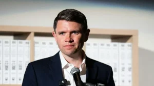 A man in a suit stands in front of a backdrop of binders, holding a microphone, suggesting he is giving a speech or presentation.