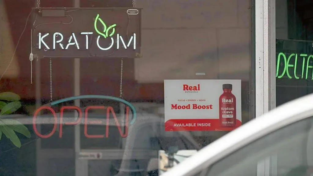 The image shows a storefront window with a neon sign that reads "KRATOM" and an "OPEN" sign, along with a promotional poster for "Real Mood Boost" featuring Kratom and Kava.