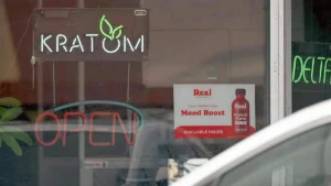 The image shows a storefront window with a neon sign that reads "KRATOM" and an "OPEN" sign, along with a promotional poster for "Real Mood Boost" featuring Kratom and Kava.