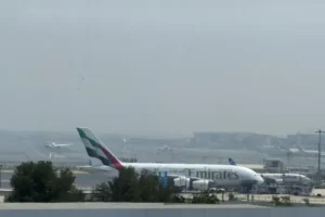 An Emirates airplane is parked on the tarmac at an airport, with other aircraft and airport infrastructure visible in the background.