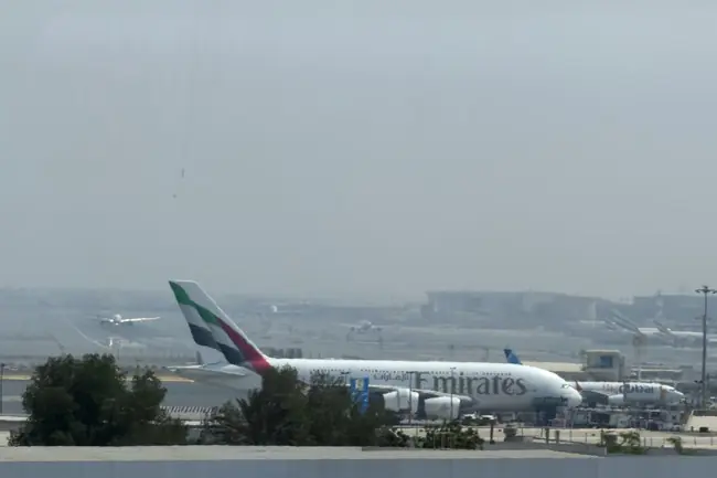 An Emirates airplane is parked on the tarmac at an airport, with other aircraft and airport infrastructure visible in the background.
