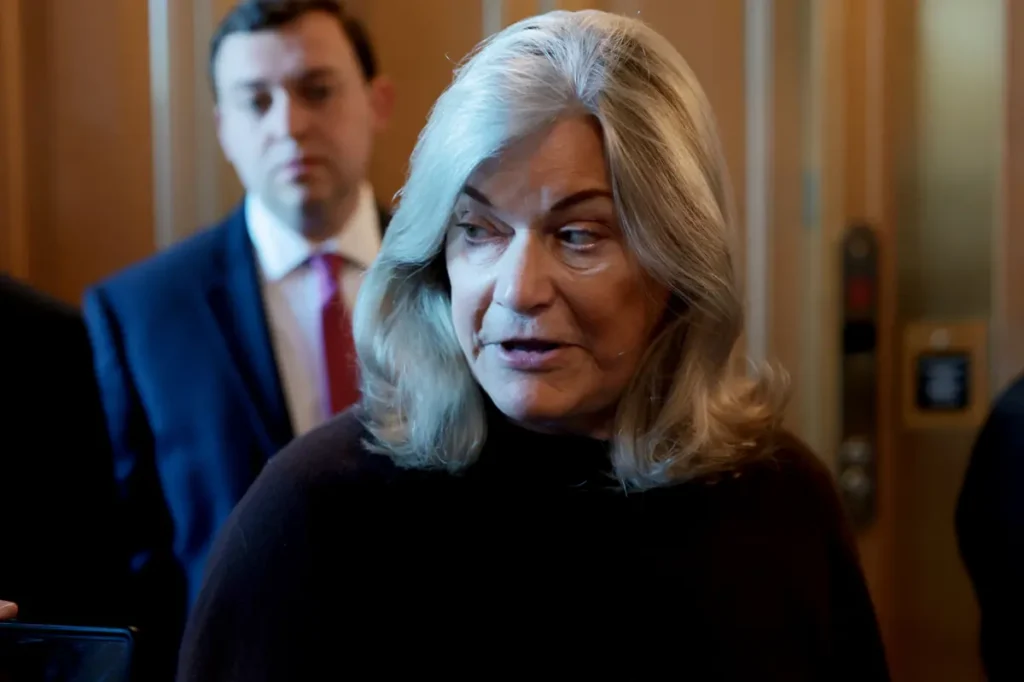 A woman with gray hair is speaking in what appears to be a formal setting, with a man in a suit standing behind her.