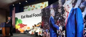 A man speaks at a podium on a stage with a backdrop that reads "Eat Real Food," while a large screen behind him shows a handshake between two individuals.