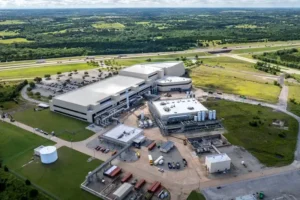 The image shows an aerial view of a large industrial facility surrounded by greenery and a highway in the background.