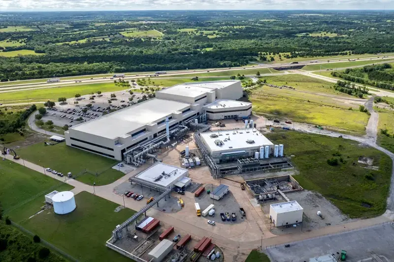 The image shows an aerial view of a large industrial facility surrounded by greenery and a highway in the background.