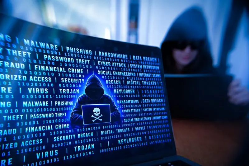 The image depicts a hooded figure holding a laptop with a skull and crossbones symbol, surrounded by a backdrop of binary code and cybersecurity-related terms.