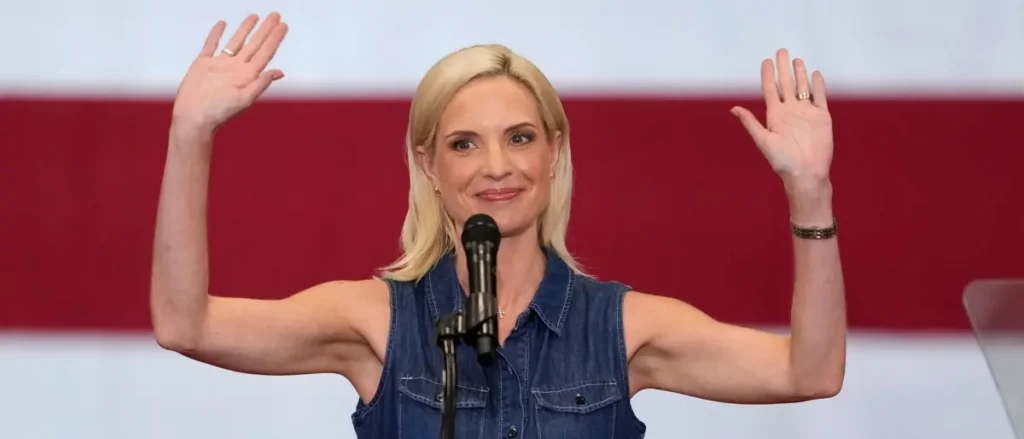 A woman in a denim shirt raises her hands in a gesture of greeting or acknowledgment while speaking into a microphone.