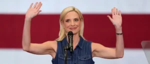 A woman in a denim shirt raises her hands in a gesture of greeting or acknowledgment while speaking into a microphone.