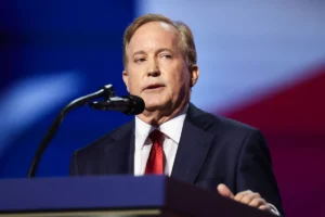 A man in a suit and red tie is speaking at a podium in front of a blurred background.