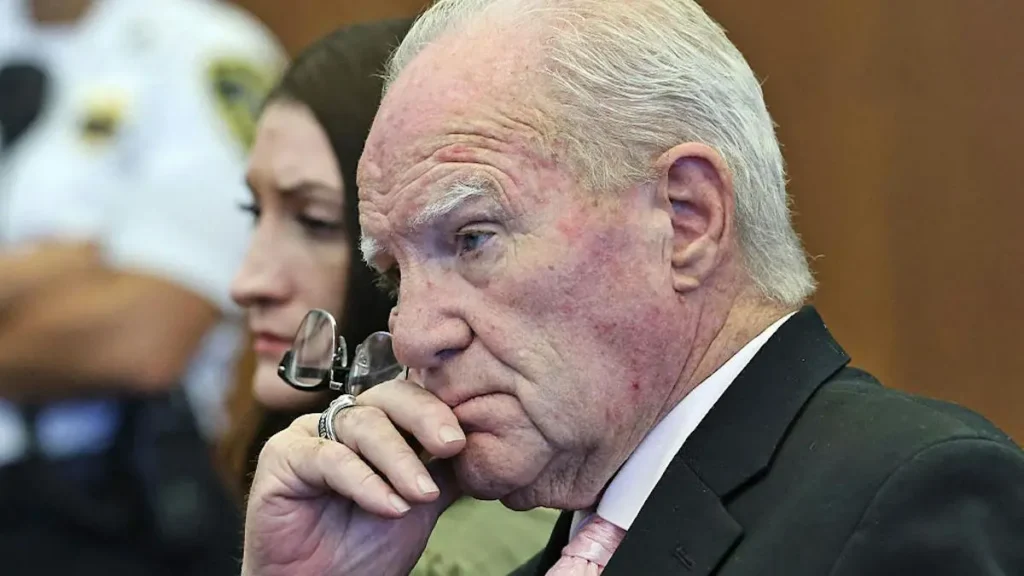 The image shows a man in a suit and tie, appearing contemplative, with a woman and another person in the background.
