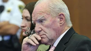 The image shows a man in a suit and tie, appearing contemplative, with a woman and another person in the background.