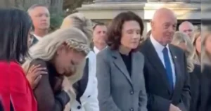 A group of people, including a woman in a white headscarf, are gathered in what appears to be a formal or somber event.