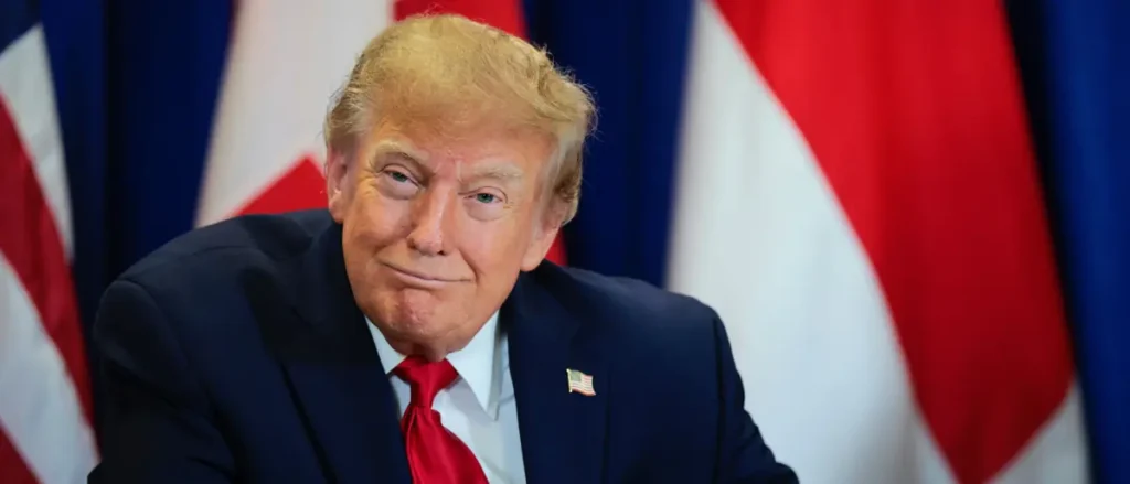 The image shows a person in a suit with an American flag pin, seated in front of multiple flags, suggesting a formal or diplomatic setting.