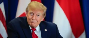 The image shows a person in a suit with an American flag pin, seated in front of multiple flags, suggesting a formal or diplomatic setting.