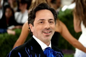 The image shows a man in formal attire, including a black suit with blue embellishments and a blue bow tie, smiling at the camera.