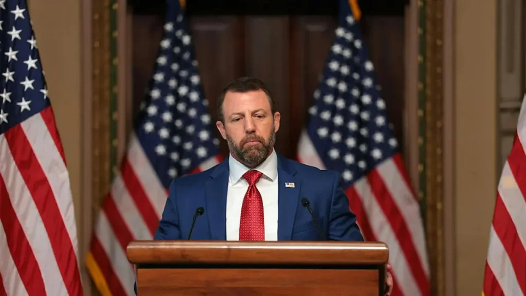 A man in a suit stands at a podium with American flags in the background, suggesting a formal or political setting.