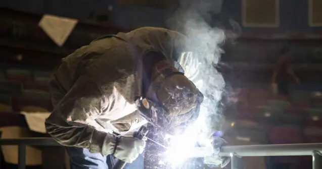 A welder is working in a workshop, surrounded by bright sparks and smoke.