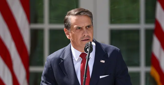 A man in a suit and tie stands at a podium with American flags in the background, delivering a speech.
