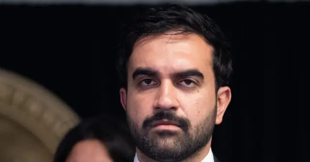 The image shows a man with a beard and short hair, dressed in formal attire, seated in a dimly lit environment.