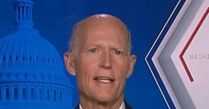 The image shows a man speaking in front of a backdrop featuring the U.S. Capitol building and the word "WASHINGTON."