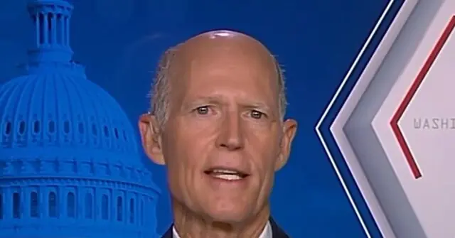 The image shows a man speaking in front of a backdrop featuring the U.S. Capitol building and the word "WASHINGTON."
