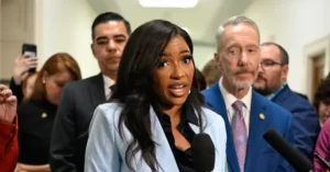 A woman speaks into a microphone in front of a group of people, with one person taking a photo in the background.